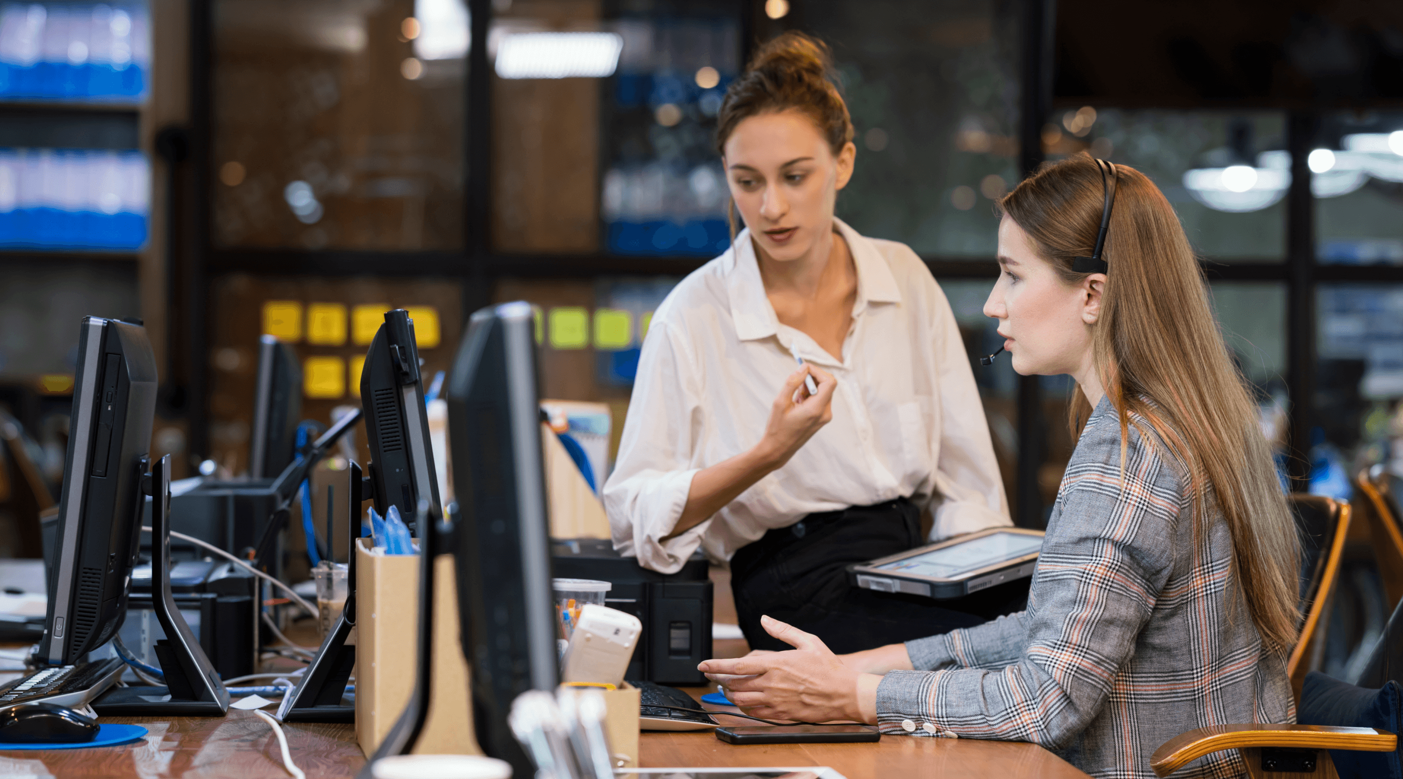 Two women talking over a desktop  and looking at a computer screen.