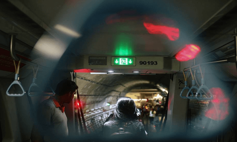 View from inside a train, looking through an open door into a dark tunnel, with a green exit sign, two people, and prominent red lens flares.