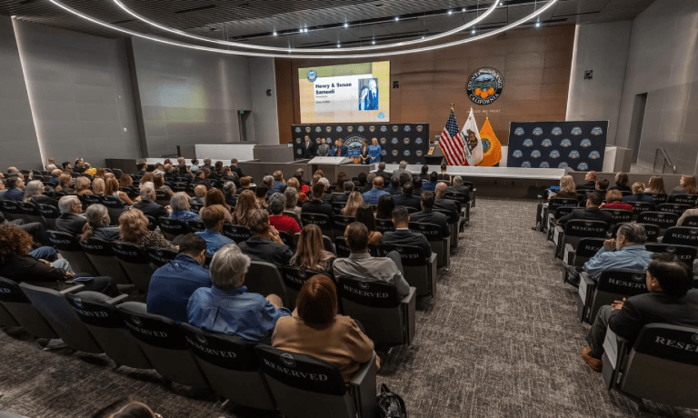 Audience in a modern auditorium facing a stage with flags and a screen displaying "Henry & Susan Samueli".